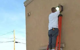 Electrician up a ladder servicing a wall light fixture on the side of a commercial building