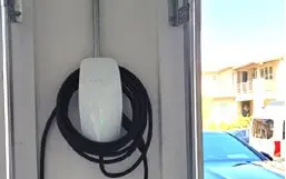 Wall charger installed inside a garage with its cable hung on the hook and a car parked outside