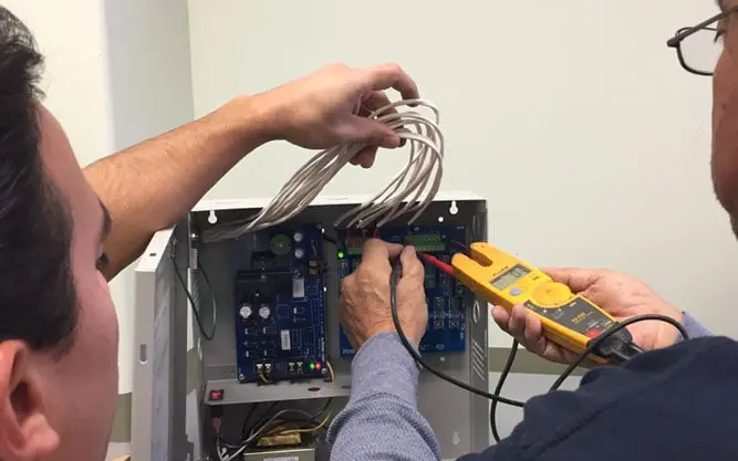 Two electricians testing an open security system panel, one holding a clamp meter against the board